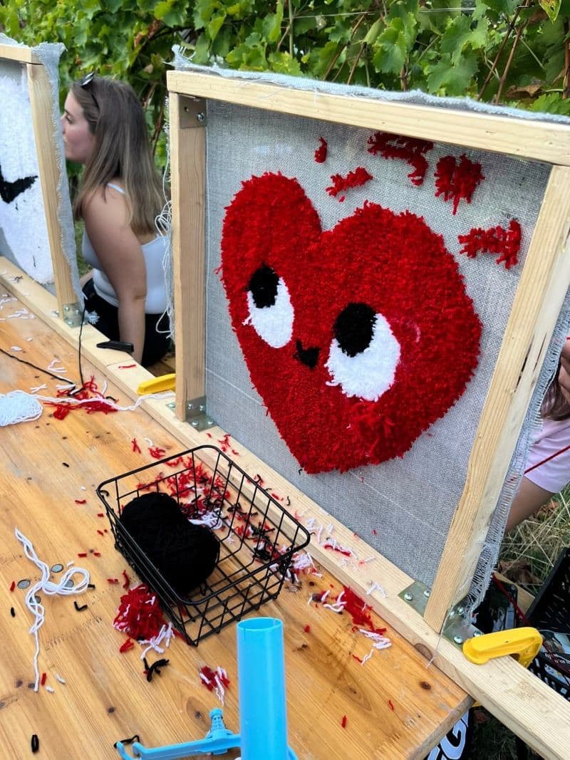 A red heart-shaped tufted rug on a wooden frame, with yarn and a tufting gun on the table