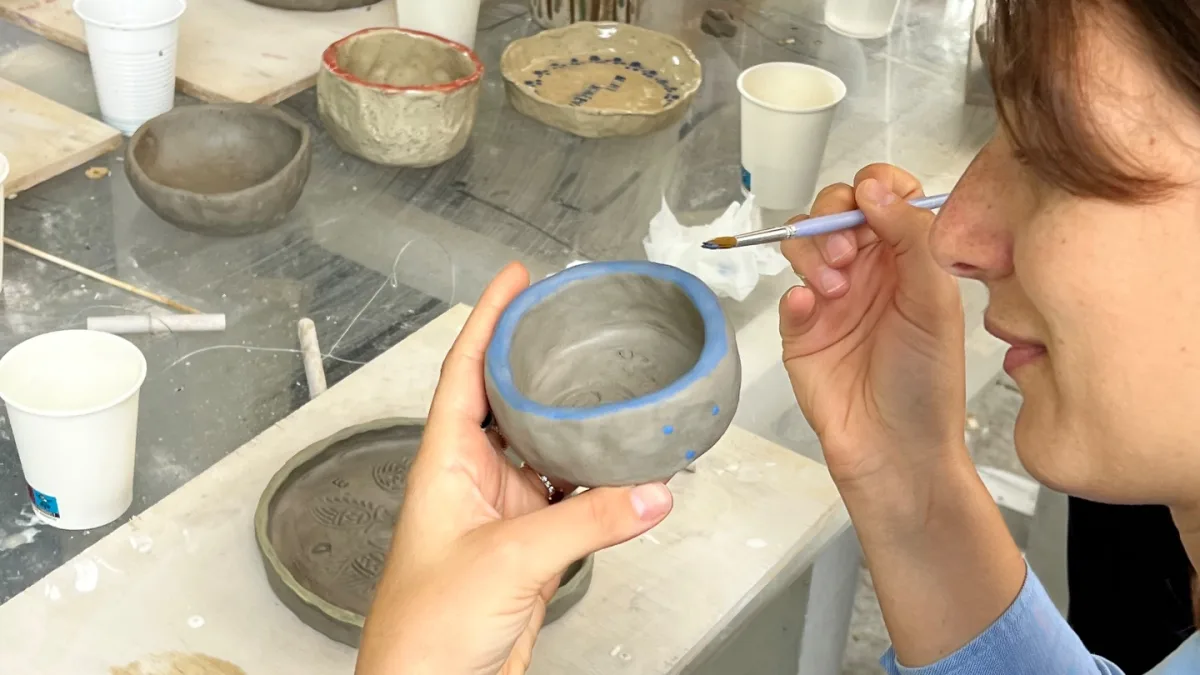 Una persona rifinisce a mano il bordo di una ciotola di ceramica su un banco da lavoro, con altri pezzi crudi accanto